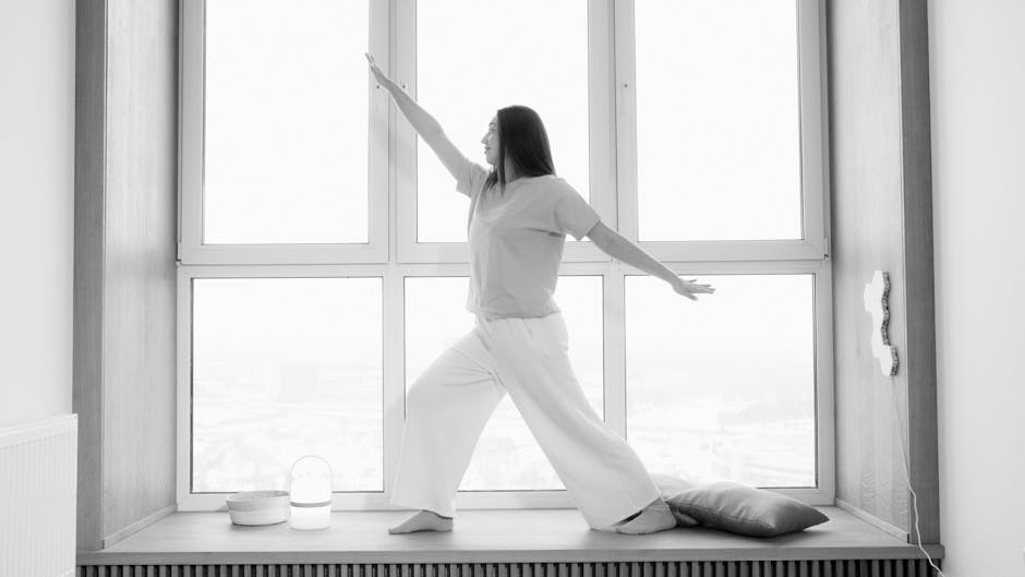 Woman in a white top performing a peaceful yoga pose next to a large window at twilight.
